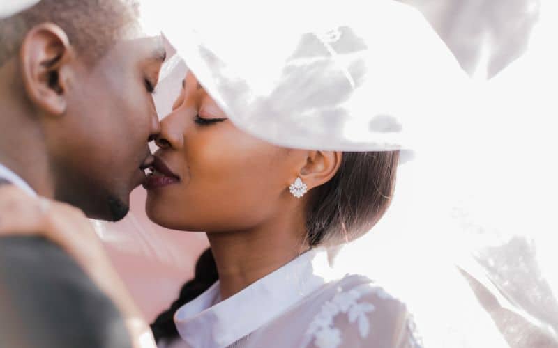 A close-up of a newlywed couple sharing a tender kiss, with the bride's veil softly draped over them. The bride wears elegant floral earrings and has her eyes gently closed, creating an intimate and romantic moment.