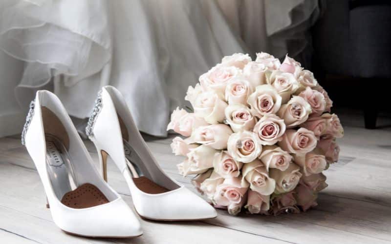 A pair of elegant white high-heeled shoes is placed next to a bouquet of pale pink roses on a wooden floor. In the background, part of a wedding dress can be seen, adding to the bridal theme of the image.