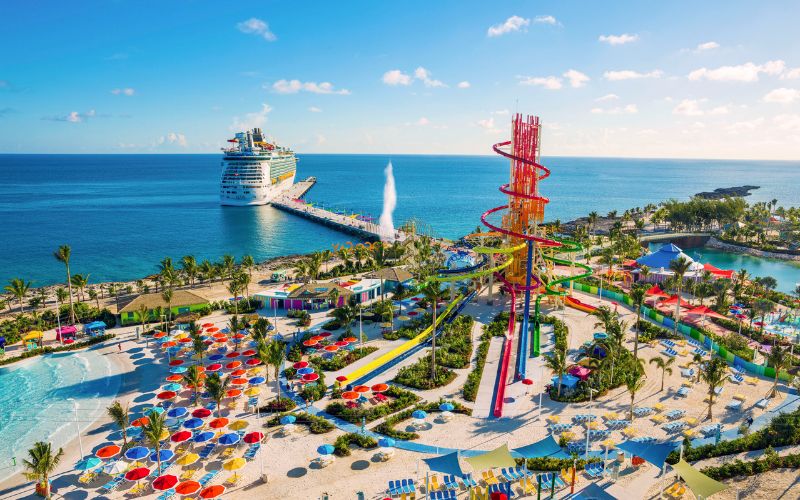 This vibrant aerial image showcases Perfect Day at CocoCay, Royal Caribbean's private island destination in the Bahamas. The photo captures the island's extensive waterpark, featuring colorful waterslides, including a massive red spiral slide, a wave pool, and an expansive swimming area. A large cruise ship is docked at the island's pier, adding to the scene's dynamic and festive atmosphere. The island is surrounded by lush greenery and crystal-clear turquoise waters, creating a perfect tropical escape for visitors. The bright colors and fun-filled water activities highlight the island as a family-friendly vacation hotspot.
