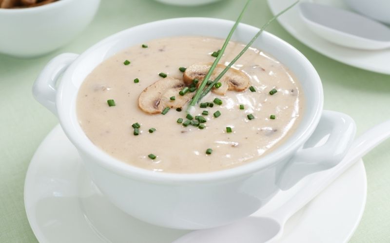 The image shows a bowl of creamy mushroom soup garnished with chopped chives and a few mushroom slices on top. The soup has a smooth, rich texture, served in a white bowl with a matching plate and spoon, creating an elegant presentation. The subtle green background complements the fresh garnish, enhancing the appetizing look of the dish.