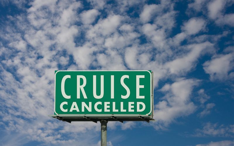 Road sign against a bright blue sky dotted with fluffy clouds reads 'CRUISE CANCELLED' in bold white letters, conveying a clear message about a change in travel plans.