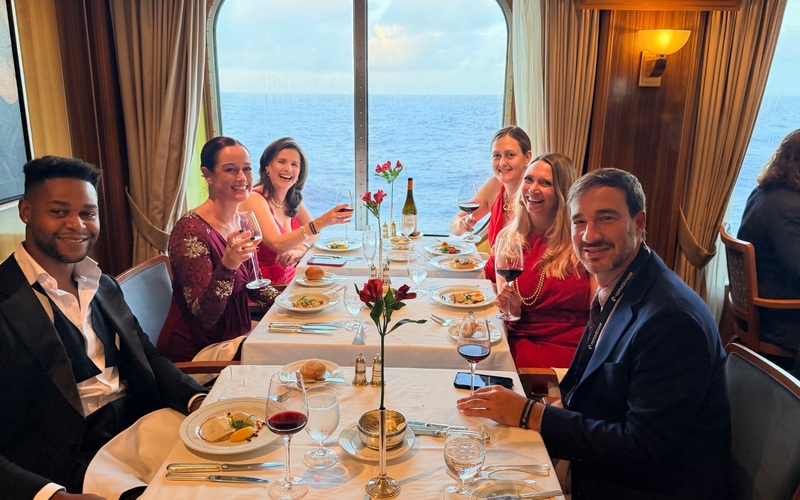 Six diners smiling around a dining table beside windows overlooking the sea.