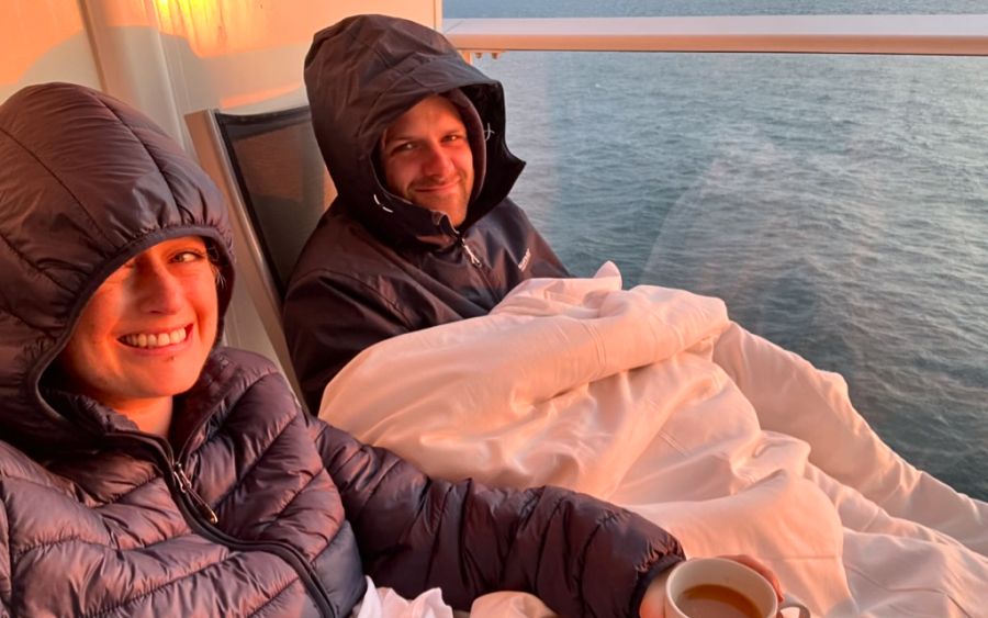 Cruise Mummy and her husband are seated on a cruise ship balcony, wrapped in blankets and wearing hooded jackets to stay warm. They are smiling and enjoying a cozy moment together, with the ocean visible in the background as the sun sets, casting a warm glow on their faces.