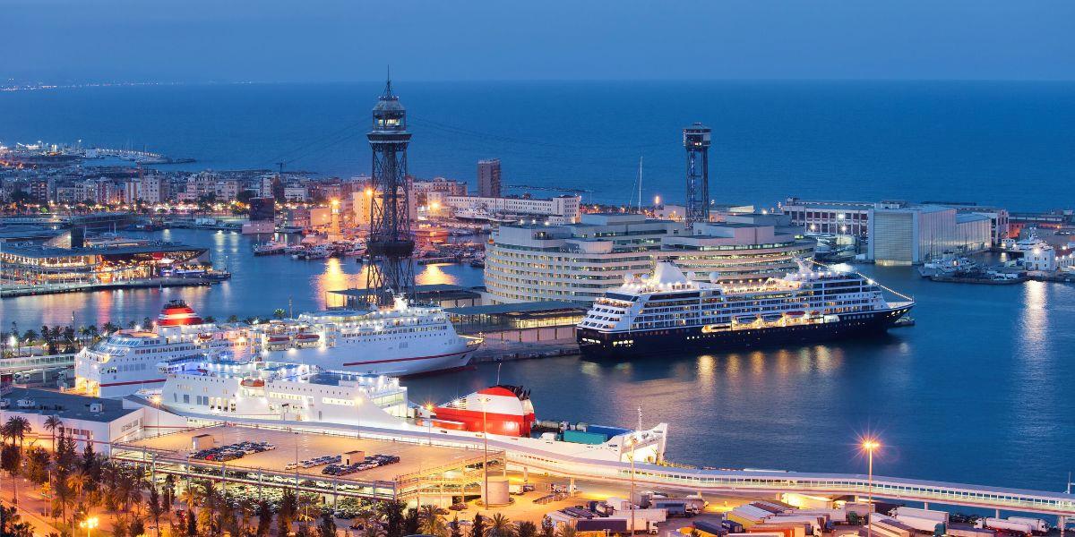 A bustling cruise port terminal at night, beautifully illuminated against the evening sky. The scene is in Barcelona, known for its iconic Port Vell. Several cruise ships are docked at the terminal, with their lights reflecting on the calm waters of the harbor. The image includes the famous Columbus Monument and the distinctive cable car tower, adding a cultural and historic dimension to the modern, lively maritime setting. The city's skyline in the background, lit under the night sky, adds a vibrant backdrop to this active port, capturing the essence of a major European cruise hub.