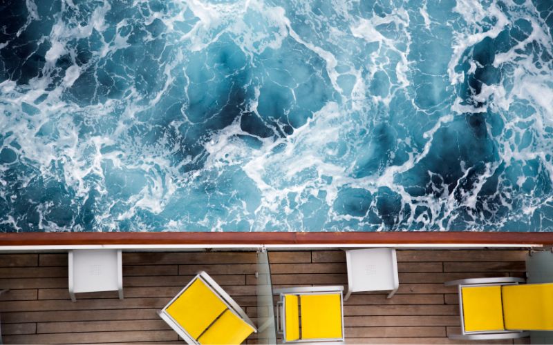 The image shows a cruise ship deck with yellow lounge chairs and tables, positioned alongside a railing overlooking a turbulent, frothy sea. The churning, deep blue water suggests stormy or rough weather, contrasting with the bright, orderly arrangement of deck furniture. This top-down perspective captures the power of the ocean and the contrast between the natural forces and the ship's calm deck setup.