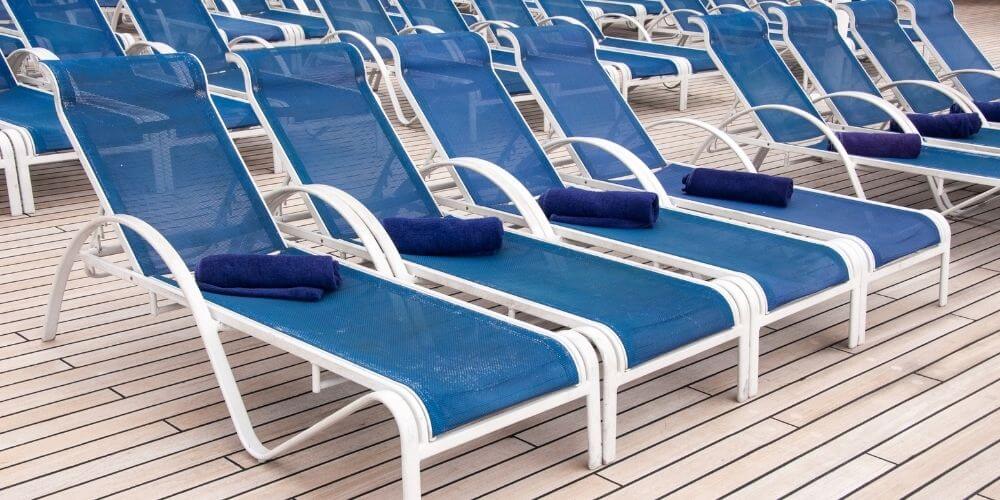 A row of blue lounge chairs on a cruise ship deck, each with a neatly folded blue towel. The wooden deck adds to the clean and organized appearance, ready for guests to relax and enjoy the sun.