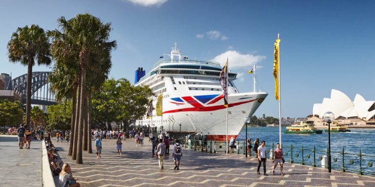 Cruise ship passengers are disembarking at a port in Sydney, with the iconic Sydney Opera House and Sydney Harbour Bridge visible in the background. The scene is bustling with people walking along the waterfront promenade, lined with palm trees and flags. The large cruise ship, adorned with a Union Jack design on its bow, is docked, creating a vibrant and picturesque setting.