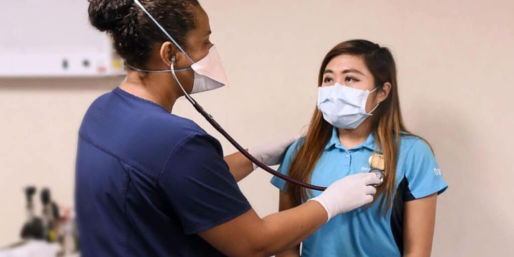 A nurse on a cruise ship, wearing a blue medical uniform and a mask, is using a stethoscope to check the heartbeat of a young woman in a light blue uniform. Both are wearing masks, ensuring adherence to health and safety protocols. The setting appears to be a medical room on the ship, equipped for onboard healthcare.