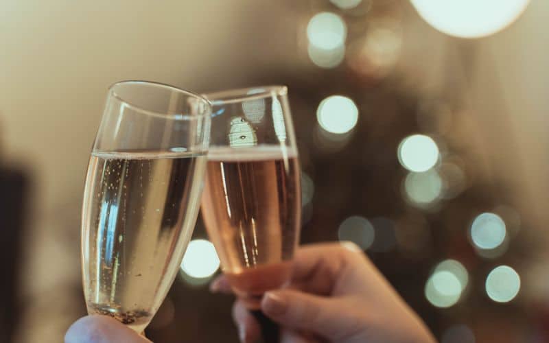 Two hands clinking champagne flutes, one with clear bubbly and the other with a pink hue, in a celebratory toast. The background is softly blurred, with hints of a festive Christmas tree, suggesting a warm, holiday celebration.