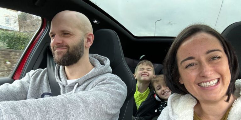 A happy family moment inside a car with a bald man in the driver's seat, two children making playful faces in the back, and a woman with a big smile taking a selfie. The car's interior suggests a comfortable and modern vehicle.