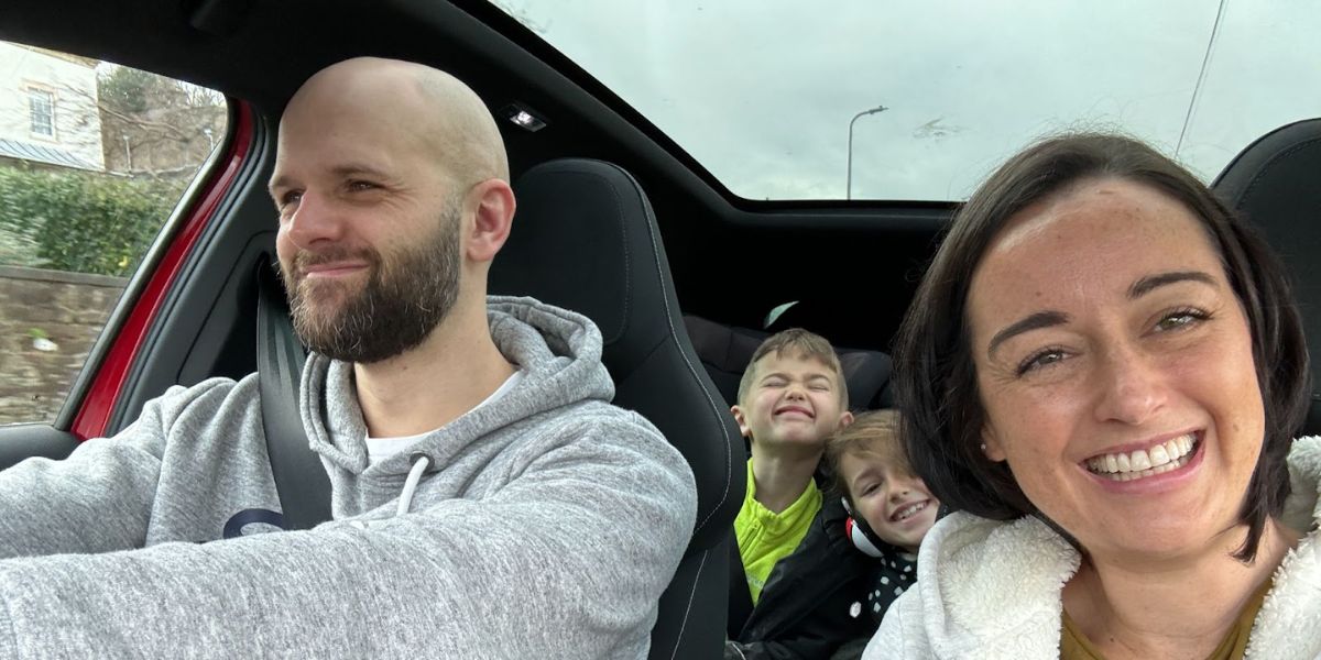 A happy family moment inside a car with a bald man in the driver's seat, two children making playful faces in the back, and a woman with a big smile taking a selfie. The car's interior suggests a comfortable and modern vehicle.