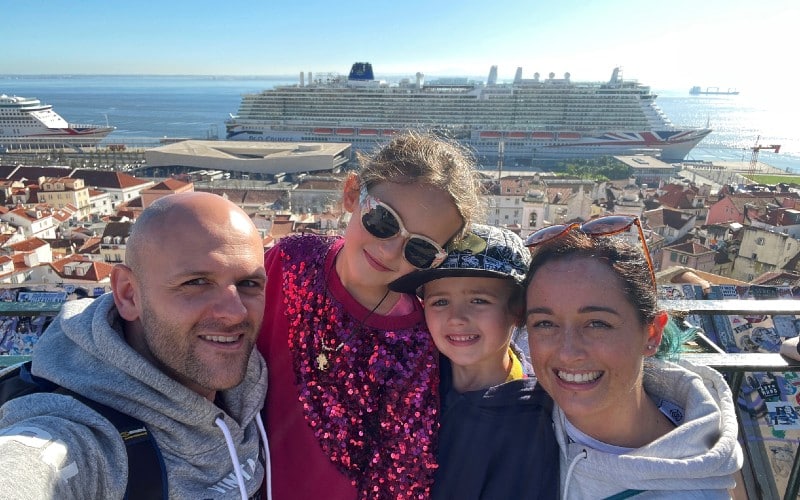 A happy family on a cruise excursion takes a smiling selfie with a picturesque view of the cityscape and docked cruise ships in the background, capturing a memorable moment of their vacation adventures.