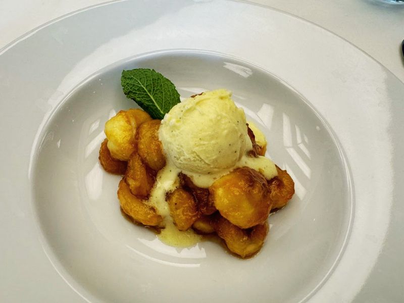 A sumptuous dessert of golden-browned puffs accompanied by a scoop of vanilla ice cream, drizzled with a creamy sauce, and garnished with a mint leaf, all presented elegantly on a white plate.