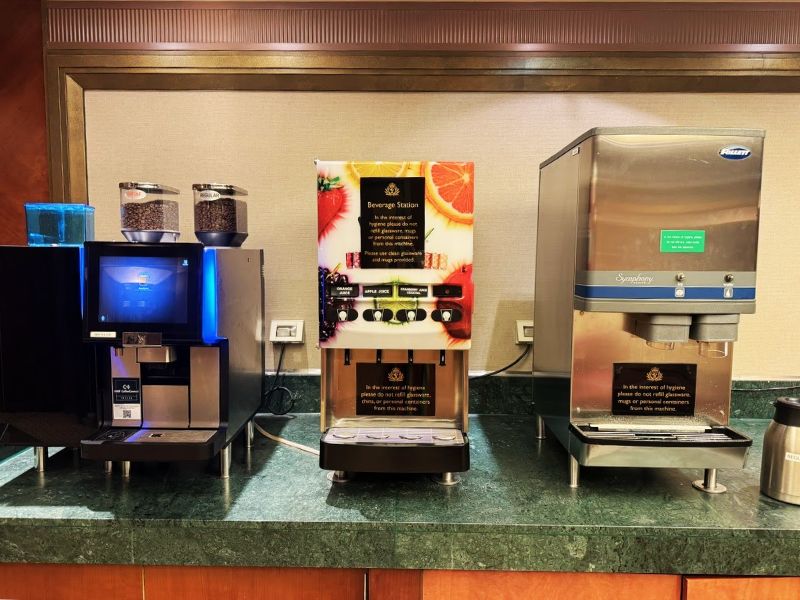 A beverage station on a Cunard cruise ship, featuring coffee, juice, and water dispensers, with signs instructing passengers to use provided cups for hygiene purposes.