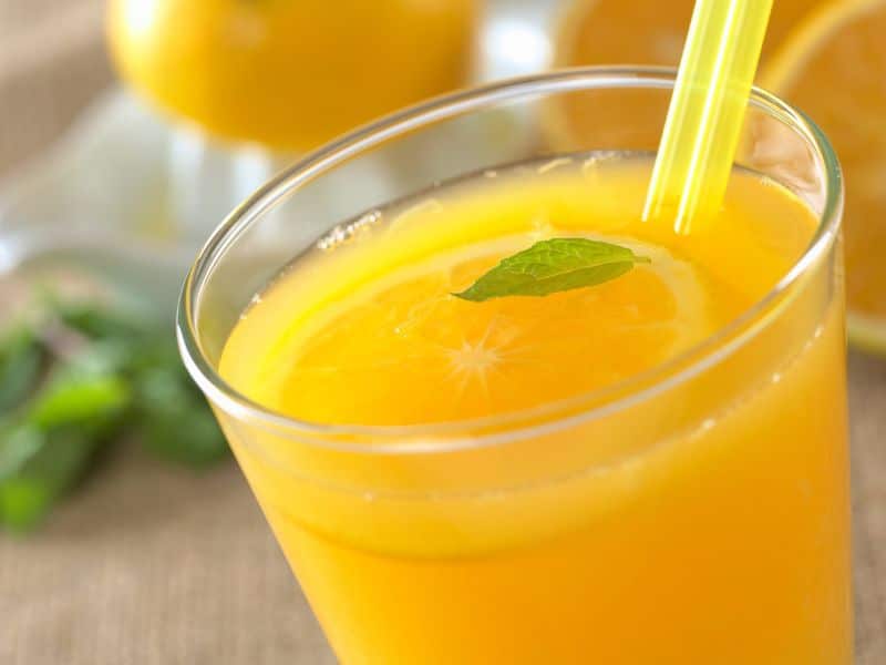 A close-up of a glass of freshly squeezed orange juice with a vibrant, refreshing appearance, garnished with a mint leaf and a slice of orange on the rim, accompanied by a straw. The background features whole oranges and a natural, soft textile, suggesting a healthy, homemade beverage.
