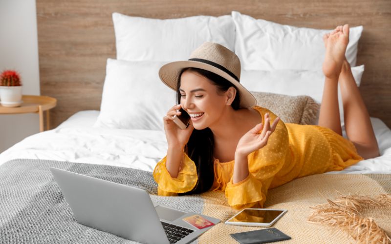 A cheerful woman in a yellow summer dress and straw hat talks on her smartphone while reclining on a bed with a laptop, tablet, and smartphone nearby, evoking a relaxed yet connected lifestyle.