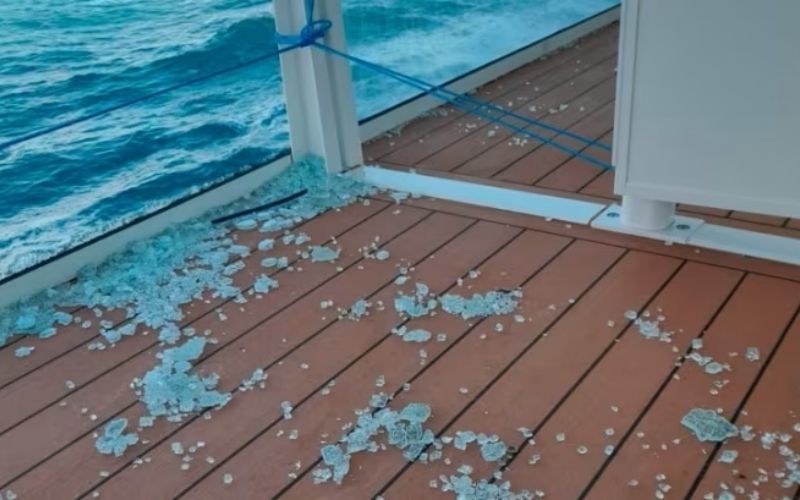 The image shows shattered glass scattered across the wooden floor of a cruise ship balcony, with pieces spread near the railing and a blue rope tied to the post. The sea is visible beyond the railing, adding to the scene of an apparent mishap on the open ocean. The broken glass sparkles against the deck, contrasting with the deep blue water in the background.