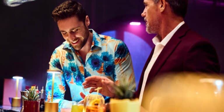 Two guests, one in a vibrant floral shirt and the other in a formal maroon jacket, share a cheerful moment at a bar on a Virgin Voyages cruise ship, reflecting the lively and stylish atmosphere on board.