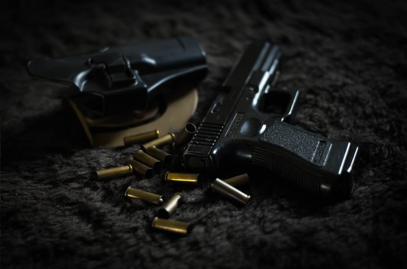 A black handgun is displayed on a dark, textured surface, surrounded by scattered bullet casings and a holster. The image emphasizes the sleek design of the firearm, with the lighting casting subtle reflections on its surface.