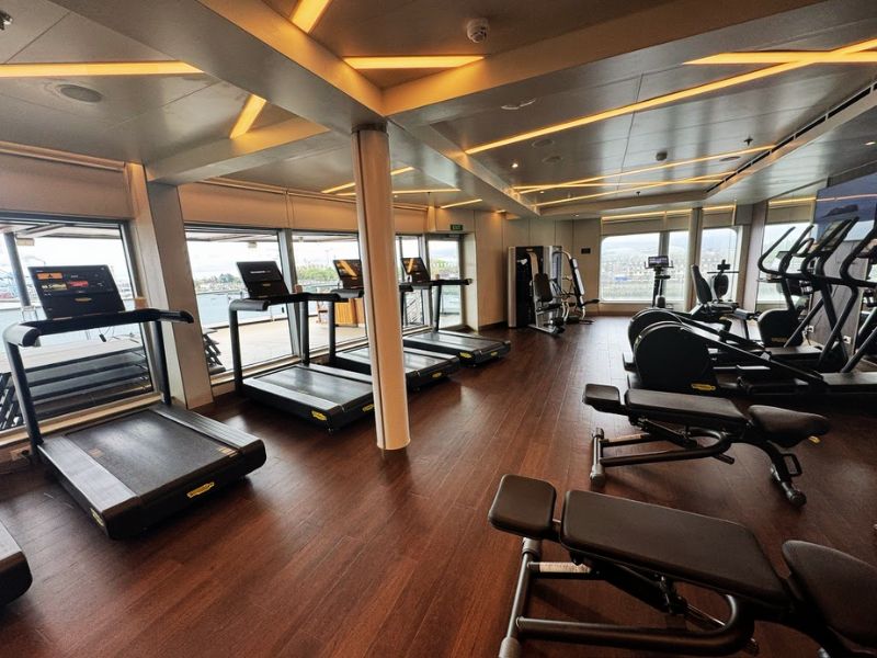 Well-equipped gym aboard the Seabourn Venture, featuring a range of modern fitness equipment. The gym is outfitted with several treadmills, elliptical machines, stationary bikes, and weight benches, all positioned to provide views through large panoramic windows. The space is bright and airy, thanks to ample natural light and a refined interior design with sleek metal columns and warm, ambient lighting. This setup invites guests to maintain their fitness routines while enjoying the scenic backdrop of their travel destinations.