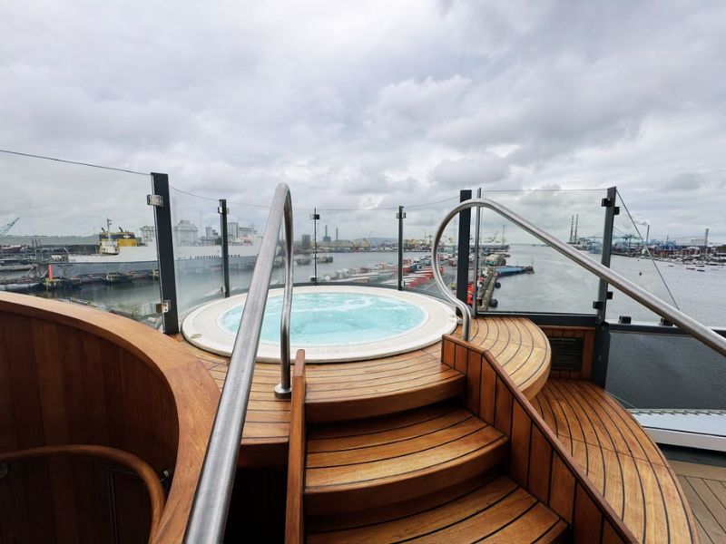 A luxurious hot tub on the deck of the Seabourn Venture, providing an inviting relaxation spot for guests. The hot tub is set within a beautifully crafted wooden structure with elegant curves and steps, facilitating easy access. Stainless steel railings add a touch of modernity and ensure safety. Positioned on the upper deck, the hot tub offers panoramic views of the port and the surrounding industrial landscape, allowing guests to unwind in warm waters while soaking in the sights. This setting perfectly combines relaxation with scenic indulgence, enhancing the cruising experience.