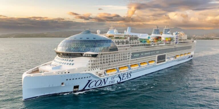 The 'Icon of the Seas' cruise ship by Royal Caribbean sails on calm seas during a vibrant sunset, with a coastal landscape in the background. This massive white ship features an array of amenities visible on the top decks, including colorful water slides and outdoor pools.