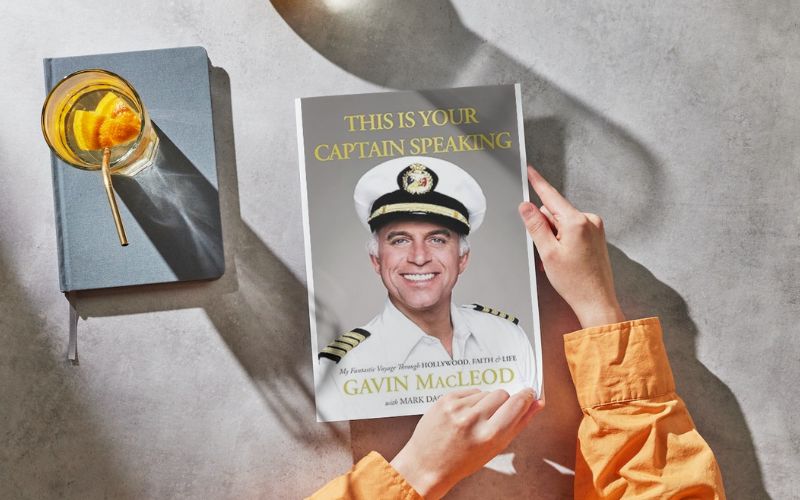Alt text: "A person's hands holding a book titled 'This is Your Captain Speaking' by Gavin MacLeod, with a portrait of the author in a captain's uniform on the cover. Next to the book is a blue journal and a glass of water with a slice of orange, all laid out on a textured surface with sunlight casting a soft shadow.