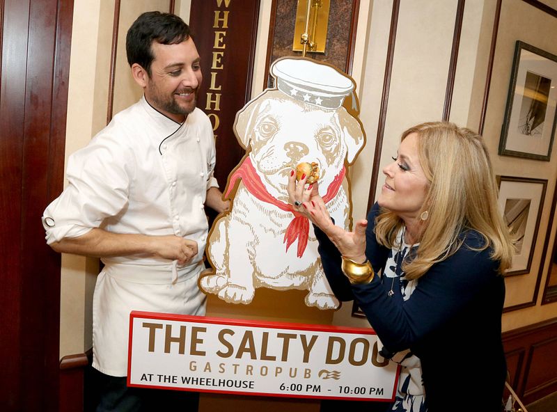 A chef in a white uniform stands next to Jil Whelan as they both touch a large illustrated sign of a bulldog wearing a sailor's hat, titled 'The Salty Dog Gastropub.' The sign also includes the pub's location and opening hours, creating a welcoming atmosphere for dining.