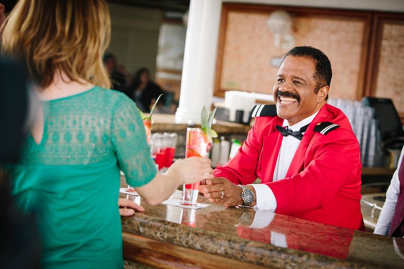 Love Boat bartender Isaac in a bright red jacket with black lapels and epaulettes engages with a person across a bar counter. He wears a bow tie and a warm, inviting smile. The bar displays an array of colorful cocktails garnished with fresh fruit, creating a lively and sociable atmosphere.