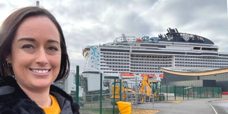 Cruise Mummy, takes a selfie with a large cruise ship featuring a distinctive blue and white design in the background, docked at a port with visible security fencing and industrial equipment.