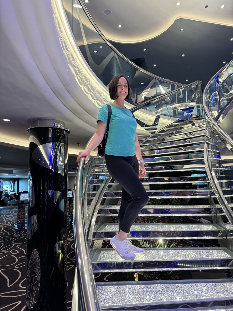 Me, standing poised on a glittering cruise ship staircase, with one hand on the railing and a smile. The luxurious setting features reflective surfaces and elegant, modern design elements, contributing to the upscale cruise ambiance.