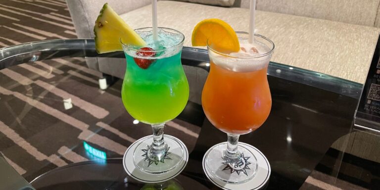 Two vibrant tropical cocktails on a glass table, with a green drink garnished with a slice of lime and a red-orange cocktail adorned with an orange slice, both served on coasters.