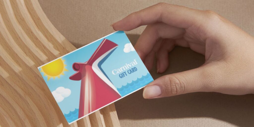 A hand is holding a 'Carnival Gift Card' against a wooden surface. The card features a stylized image of a cruise ship's bow with waves, a sun, and clouds in a blue sky, emphasizing the fun associated with a carnival cruise.