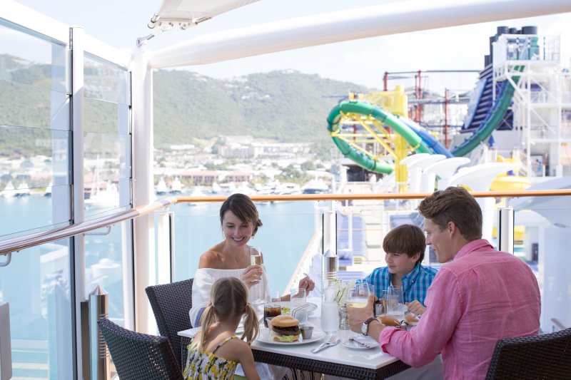 A family enjoys a meal together on NCL haven balcony with a clear view of a colorful waterslide and a picturesque port town in the background.