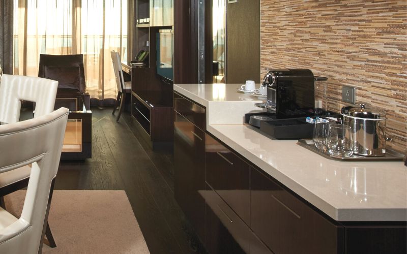 A sleek modern kitchenette in NCL haven, featuring dark wood cabinetry, a marble countertop with coffee machine and shiny stainless steel appliances, adjacent to a cozy sitting area with plush chairs.