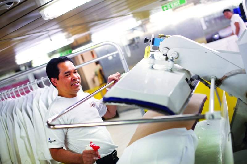 A cheerful crew member operates a large industrial ironing machine, pressing white linens in the laundry room of NCL haven, with a focus on impeccable onboard service.