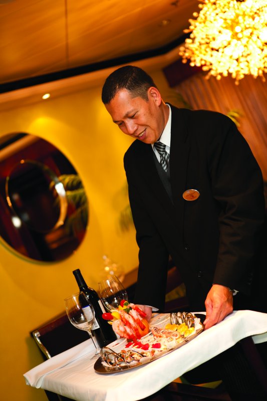 A professional waiter in a formal suit serves a seafood platter accompanied by a bottle of red wine, enhancing the fine dining experience in an elegant cruise ship restaurant.