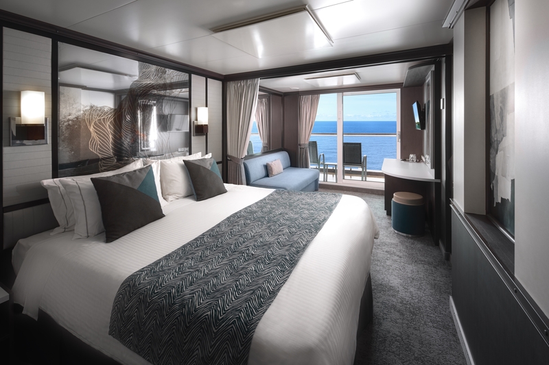 Luxury NCL Club Balcony Suites with a king-size bed adorned with a geometric-patterned throw, flanked by elegant lamps, a daybed near the panoramic window leading to a private balcony with ocean views, and a vanity area with a round stool.