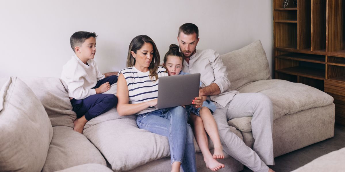 A family of four, with a mother, father, son, and daughter, are engaged with a laptop while sitting closely together on a soft beige sofa in a home setting, showcasing a moment of family connectivity and digital interaction.