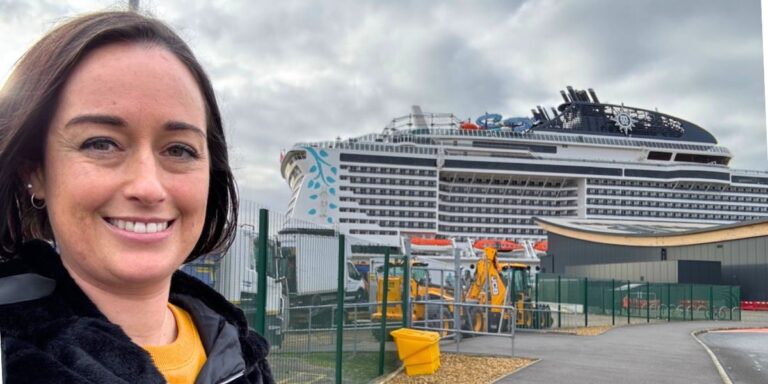 A woman smiles for a selfie with a large cruise ship docked in the background. The ship is white with multiple decks, and various construction equipment is visible in the foreground under a cloudy sky.