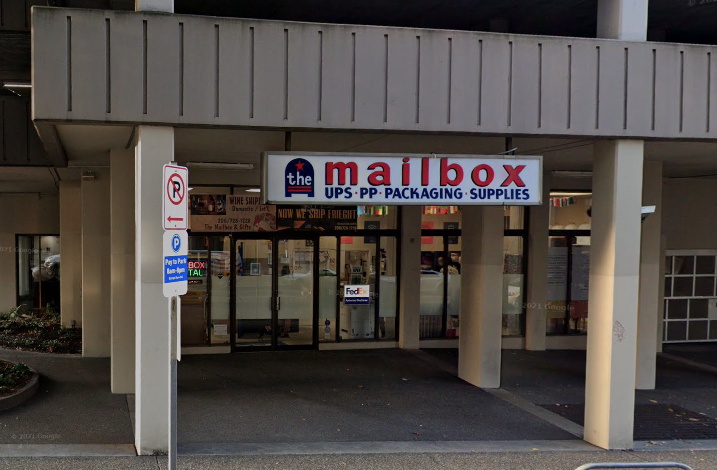 An image of 'The Mailbox' storefront, a shipping and supplies center, featuring signage for UPS, FedEx, and postal services, nestled under an overhang in a commercial area with parking signs prominently displayed.