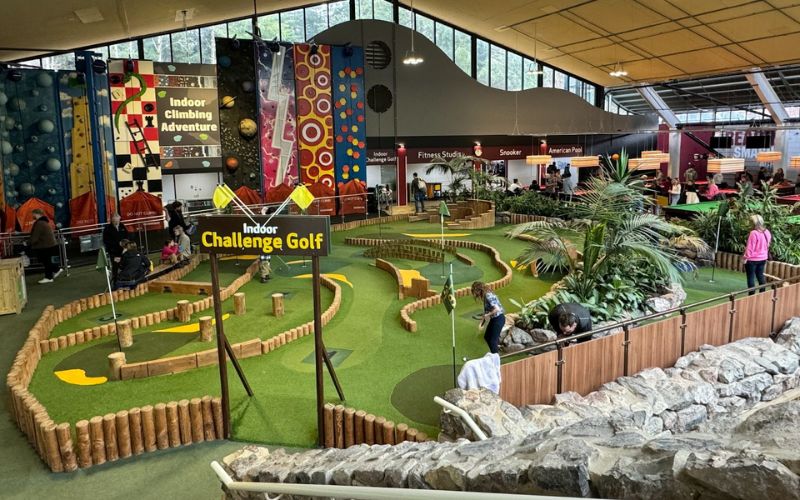 An indoor mini-golf course bustling with activity, featuring well-maintained green turf and naturalistic rock features. In the background, an indoor climbing wall and various recreational signs indicate a family-friendly sports and leisure center.