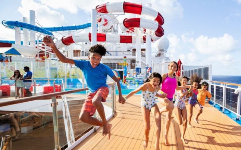 A group of joyful children racing towards the Waterworks aqua park on the deck of a Carnival Cruise ship, with clear blue skies above and the expansive ocean in the backdrop, highlighting the thrill and excitement of cruise ship activities for kids.