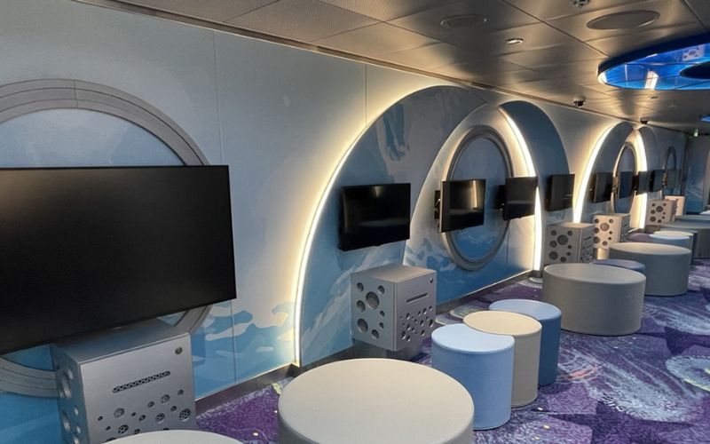 A modern gaming area designed for children, featuring multiple screens mounted on a wall with circular design elements. The room is furnished with cylindrical stools and padded seats, all set against a colorful, underwater-themed carpet. The setup is ideal for group gaming sessions and interactive entertainment.
