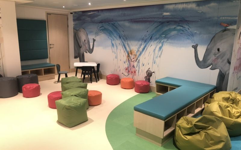 A cozy children's reading and playroom featuring colorful bean bags and cushioned benches. The walls are adorned with a whimsical mural of elephants spraying water, adding a playful touch to the room. The space is designed for comfort and creativity, with small tables and seating areas ideal for storytime and quiet play.