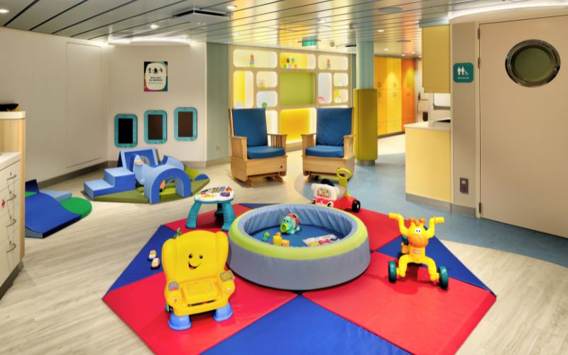 A well-equipped toddler playroom with colorful, soft play mats, a small ball pit, and various interactive toys. The room includes comfortable seating for adults and padded structures for climbing and play, creating a safe and engaging environment for young children. Bright lighting and cheerful decor enhance the inviting atmosphere.