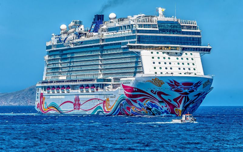 The image features a large, modern cruise ship sailing on blue ocean waters, distinguished by its vivid and colorful bow artwork. The ship is massive, multi-decked, and equipped with various amenities, as indicated by its multiple rows of balconies and advanced navigational equipment on top. A small boat can be seen nearby, emphasizing the scale of the cruise ship. The setting suggests a vibrant and luxurious travel experience.