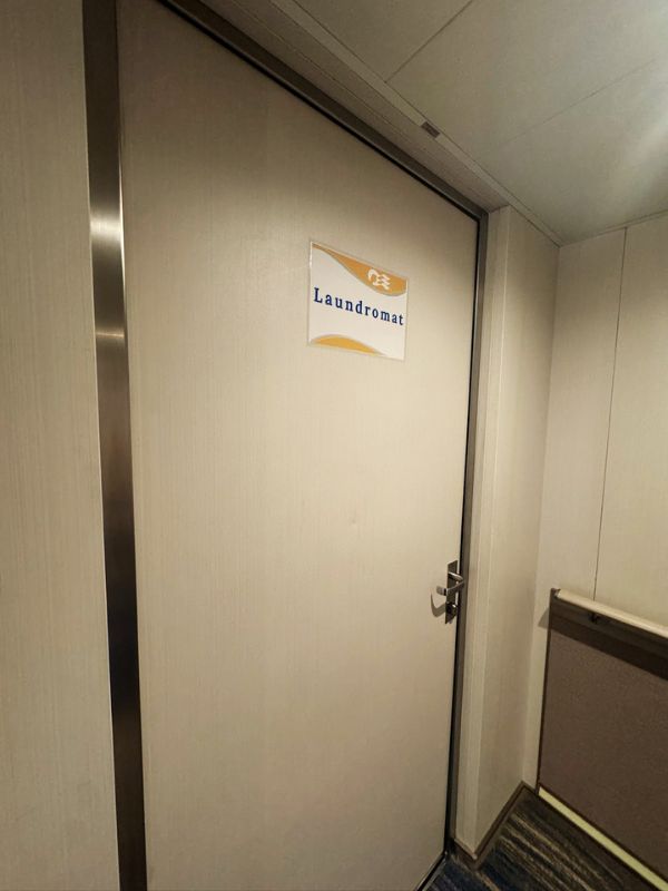 The image shows the entrance to the Laundromat on Princess Cruises' Sun Princess. The door is plain, with a simple sign that reads "Laundromat" in blue text on a white and gold background, indicating the facility's purpose. The surrounding walls are light-colored, with a minimalist design, typical of hallway or utility areas on a cruise ship. The door handle is a standard lever type, and the area appears clean and well-maintained.