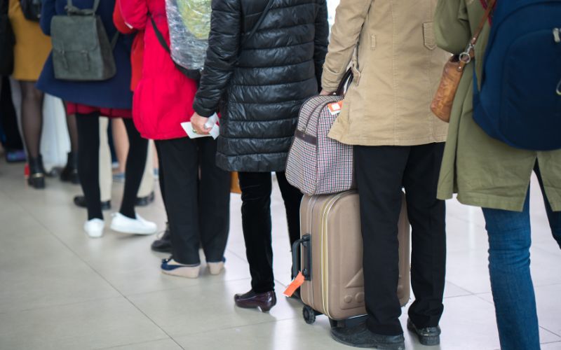 This image captures a line of passengers at a cruise terminal, each with luggage in tow. The travelers, dressed in varied seasonal attire from heavy jackets to light layers, indicate a diverse group. The focus is on the backs of the passengers, showcasing a common scene of waiting and anticipation often experienced during travel check-ins or boarding processes. This depiction highlights the busy, yet organized nature of cruise travel embarkation.