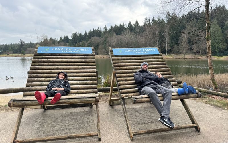 A child and an adult relaxing on large wooden loungers at Longleat Forest Center Parcs, with the serene lake and forest in the background, capturing a moment of leisure and enjoyment in the heart of nature.